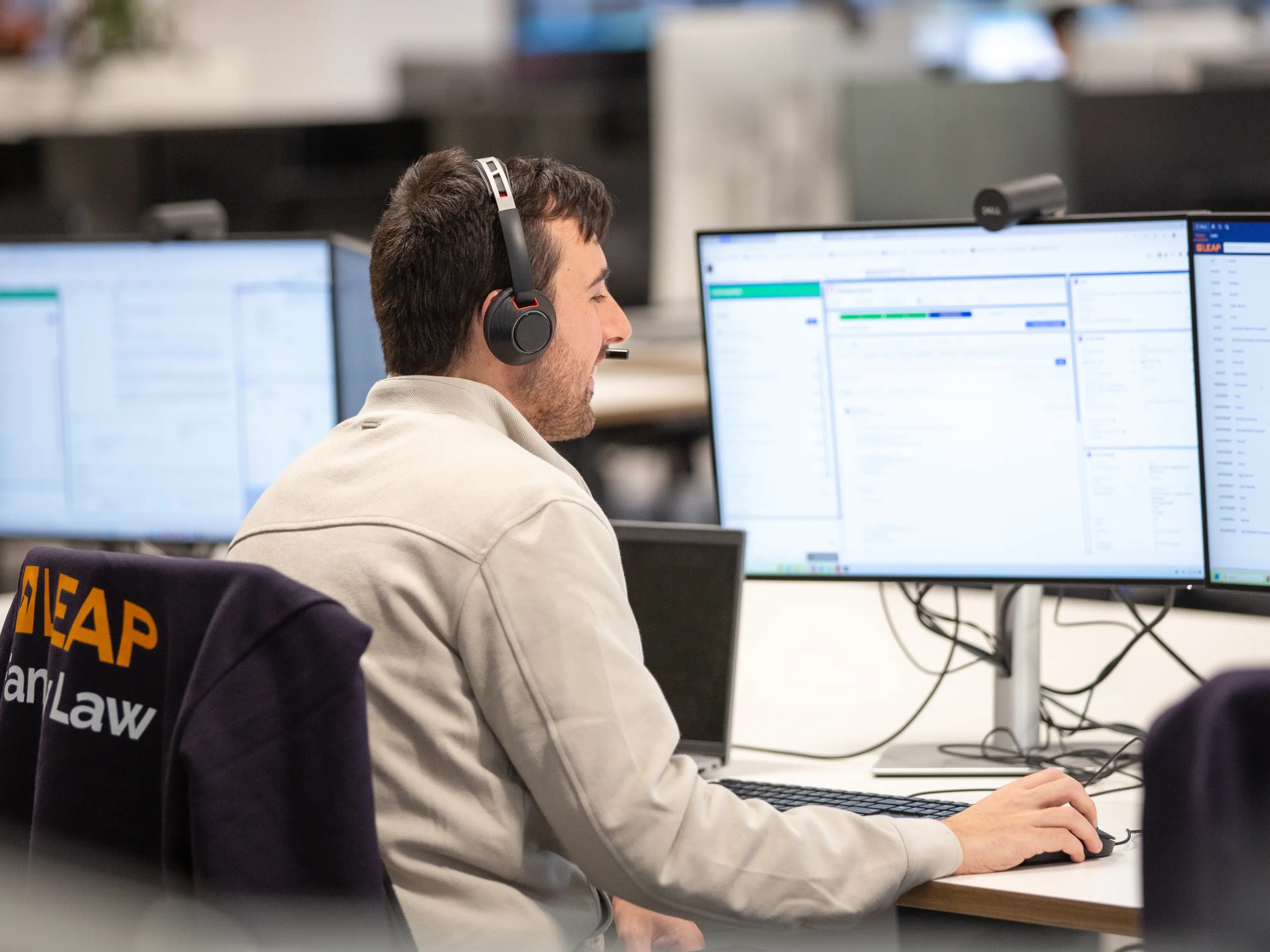 A person wearing headphones works at a desk with dual monitors displaying software applications in an office environment.
