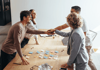Four colleagues smiling and shaking hands over a wooden table with colorful sticky notes, displaying teamwork and cooperation.