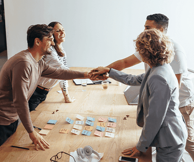 Four colleagues smiling and shaking hands over a wooden table with colorful sticky notes, displaying teamwork and cooperation.