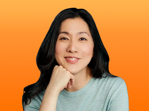 Woman with long dark hair smiling, resting her chin on her hand against an orange background.