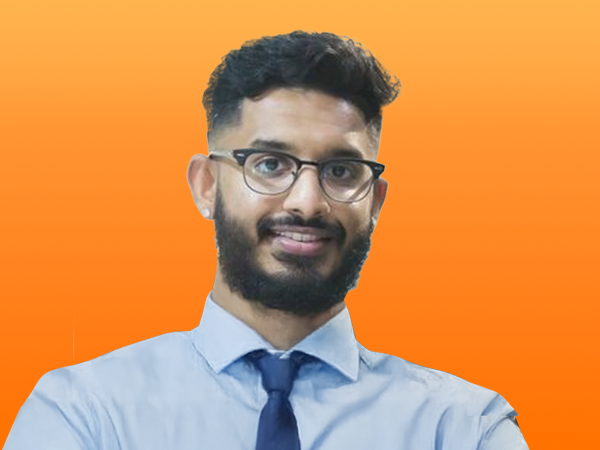 Smiling man with glasses and beard wearing a light blue shirt and navy tie against a bright orange background.