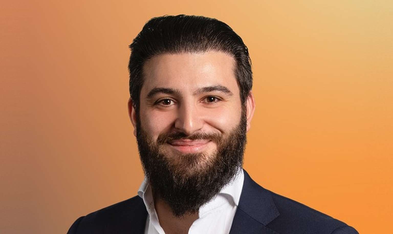 Man with dark hair and beard wearing a white shirt and dark suit, smiling against an orange gradient background.