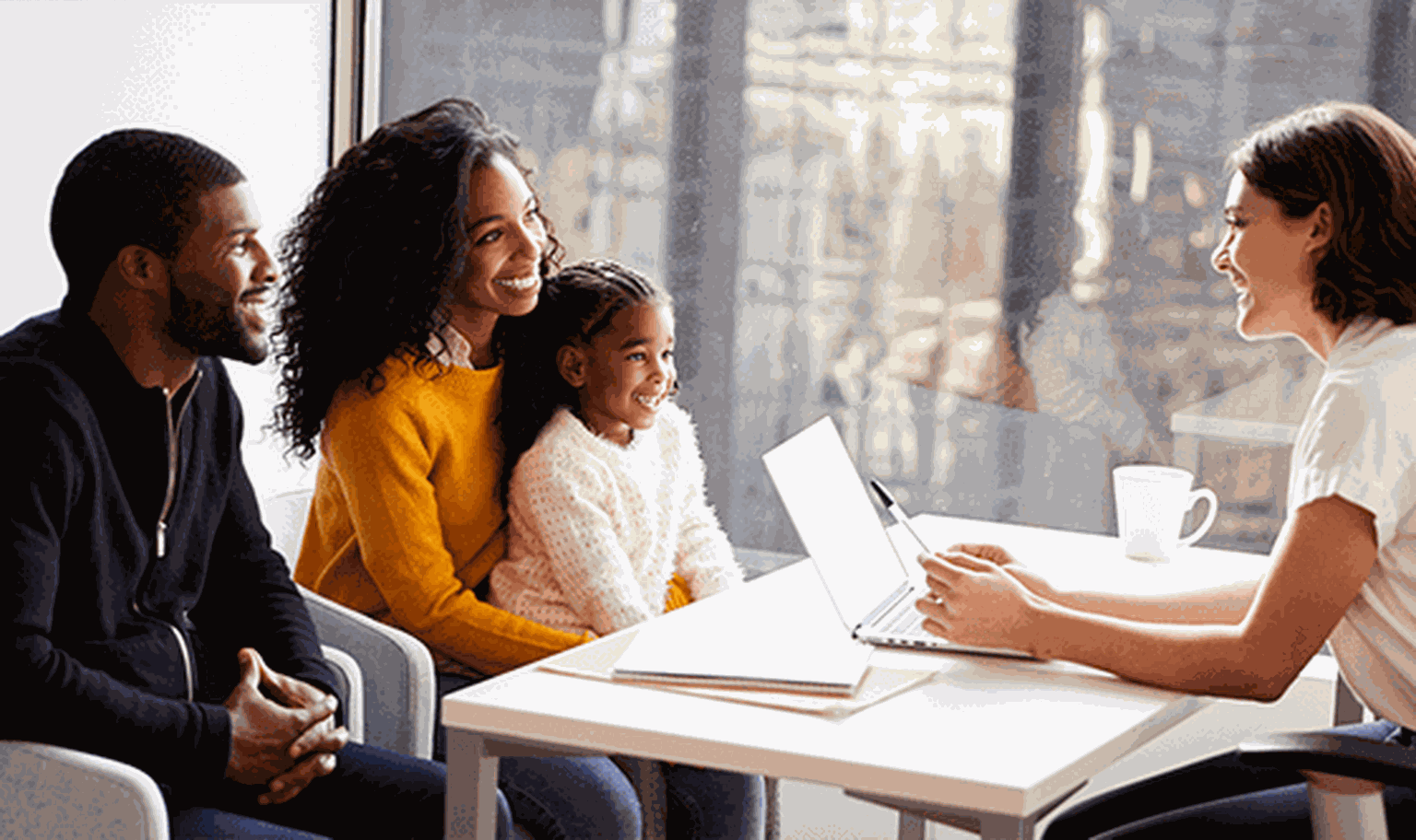A family of three smiles while talking to a woman with a laptop at a table in a bright office setting.