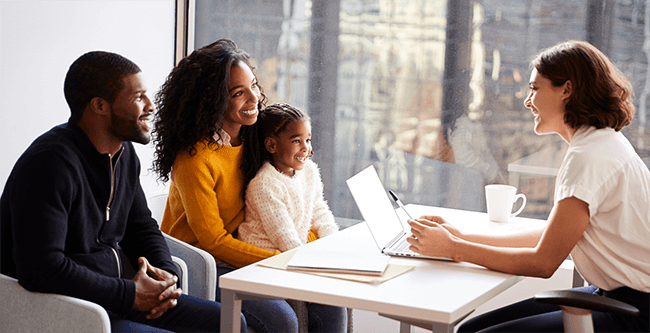 A family of three smiles while talking to a woman with a laptop at a table in a bright office setting.