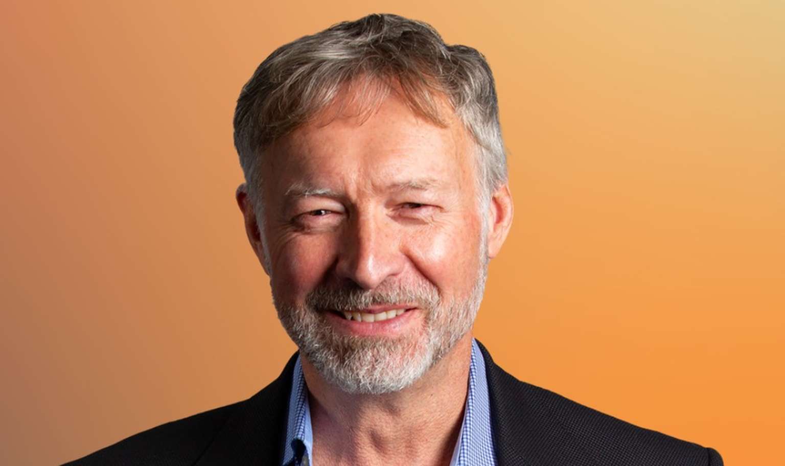 Smiling man with short gray hair and a beard, wearing a suit jacket and shirt, against an orange gradient background.