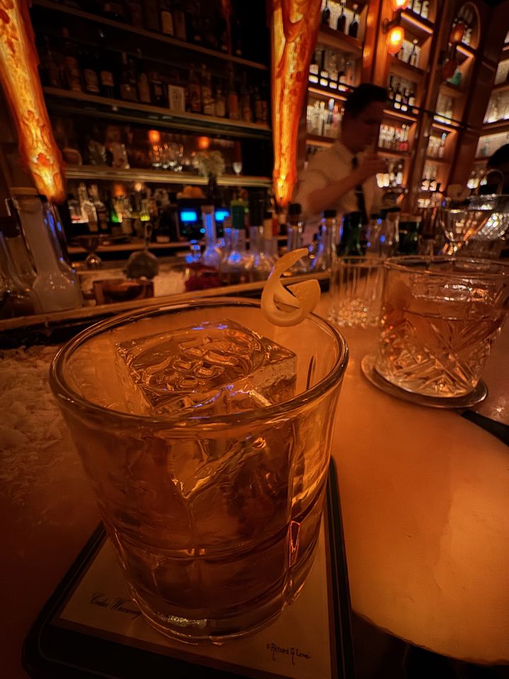 a clear amber cocktail with lemon twist garnish sitting on a candlelit bar with backlit shelves of bottles in the background