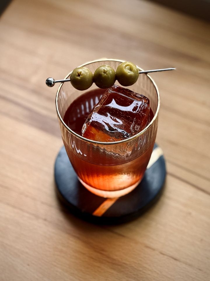 a clear red cocktail with big rock of clear ice and three speared olives as garnish sitting on a wooden table