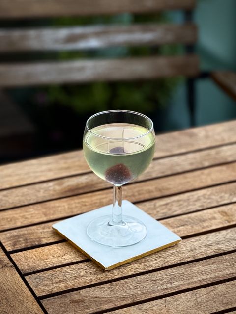 a very light green cocktail with maraschino cherry garnish in a deep coupe glass sitting on a white coaster on a wooden table