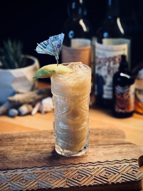 a light brown creamy cocktail with a lemon wedge and wind-blown cocktail umbrella garnish in a transparent glass tiki mug sitting on a wooden cutting board with bottles, plants, driftwood, and seashells in the background