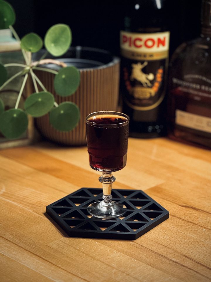 a dark red cocktail in a small coupe glass with potted plants and bottles of Amer Picon and bourbon in the background
