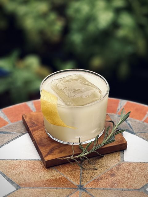 an off-white milky cocktail with big rock of clear ice, lemon twist, and lavender sprig garnish sitting on an olive wood coaster with plants in the background