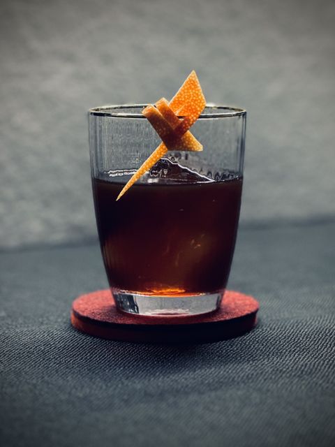 a dark brown cocktail with big rock of clear ice and orange twist garnish sitting on a maroon felt coaster against a white background