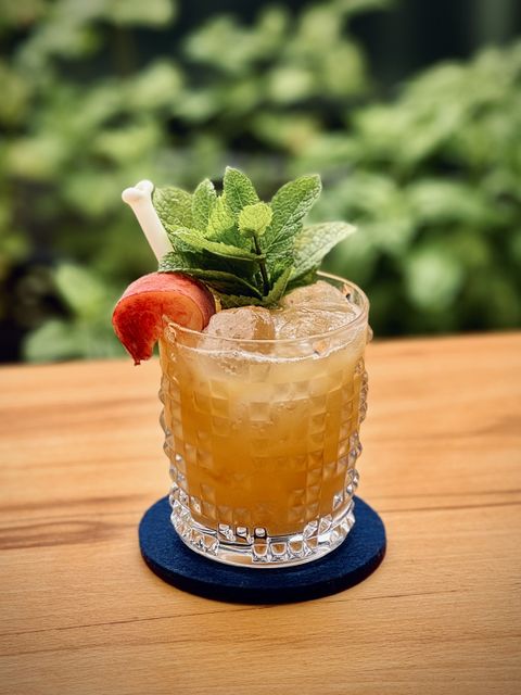 light brown cocktail with peach chunk, mint sprig and SurfSide Sips white bone straw garnish, sitting on a wooden table with mint and basil plants in the background