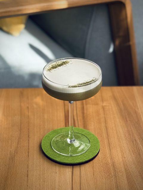 a gark green cocktail with thick white foam head and matcha tea powder garnish sitting on a green felt coaster in the morning sun