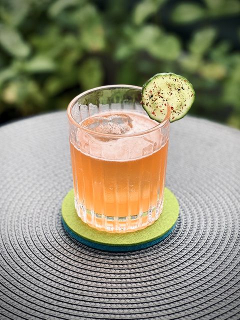 pinkish orange cocktail with big rock of clear ice and black pepper speckled cucumber slice garnish sitting on a green felt coaster with plants in the background