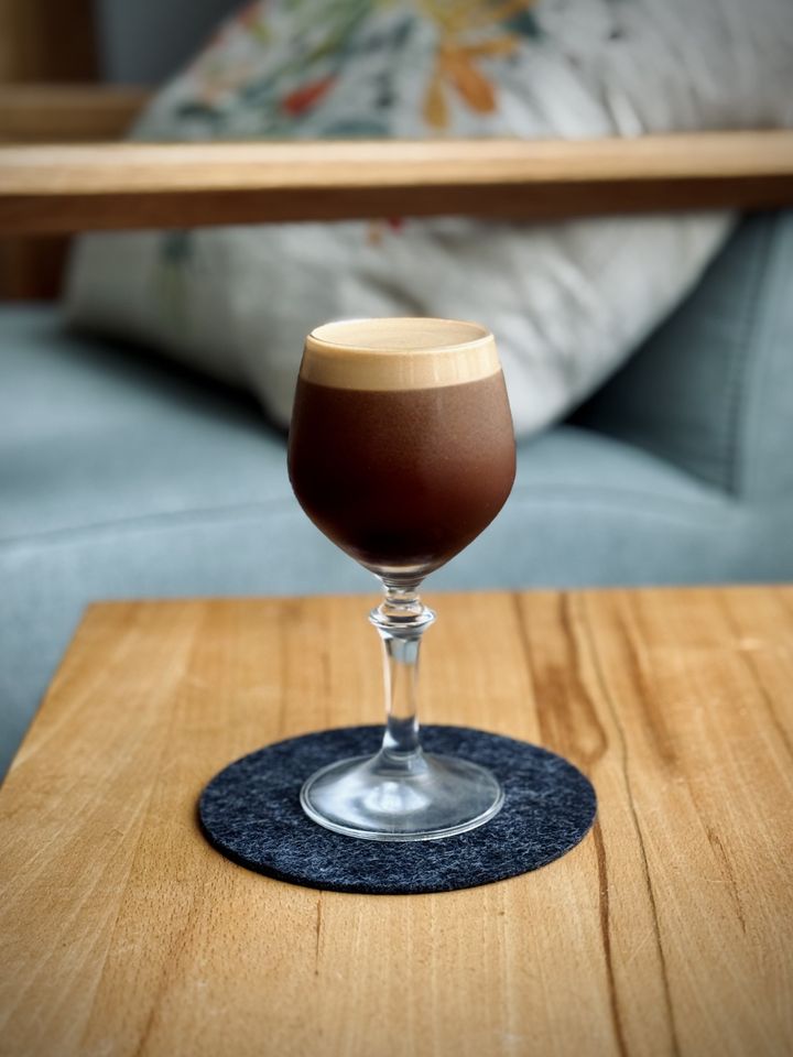 a dark brown cocktail in a coupe glass with light brown foam head. the cocktail is sitting on a black felt coaster on a wooden table.