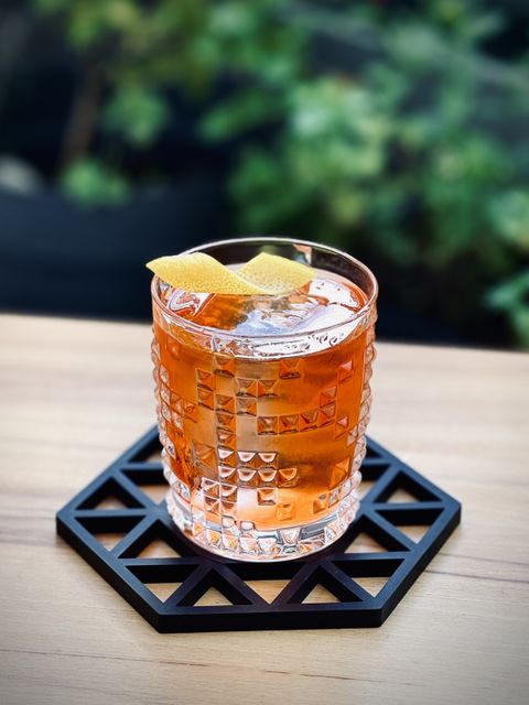 a red cocktail with big rock of clear ice and lemon peel garnish in a faceted crystal tumbler sitting on a black silicone coaster on a wooden table with plants in the background