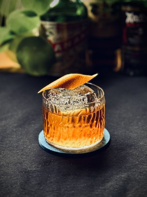 a bright amber cocktail with big rock of clear ice and orange peel garnish sitting on a black surface with bottles and plants in the background