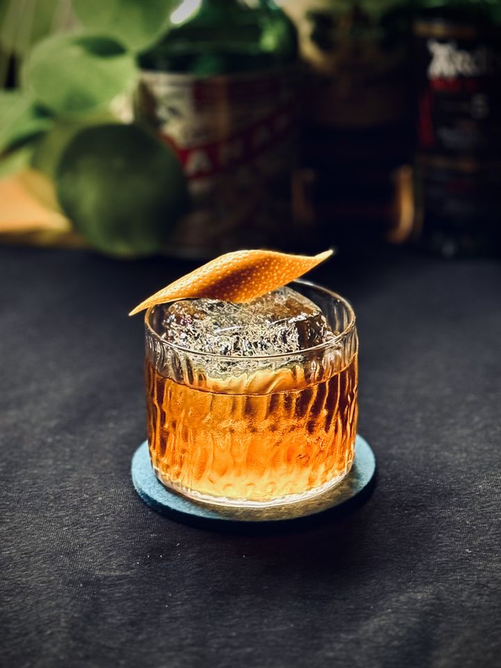 a bright amber cocktail with big rock of clear ice and orange peel garnish sitting on a black surface with bottles and plants in the background