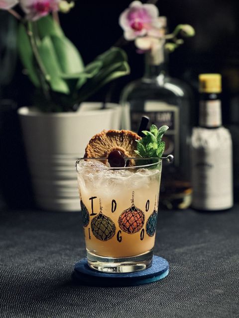 a light brown cocktail with dehydrated pineapple, maraschino cherry, and mint bouquet garnish. in the background is an orchid plant with white and pink blossoms, and a bottle of barbados rum