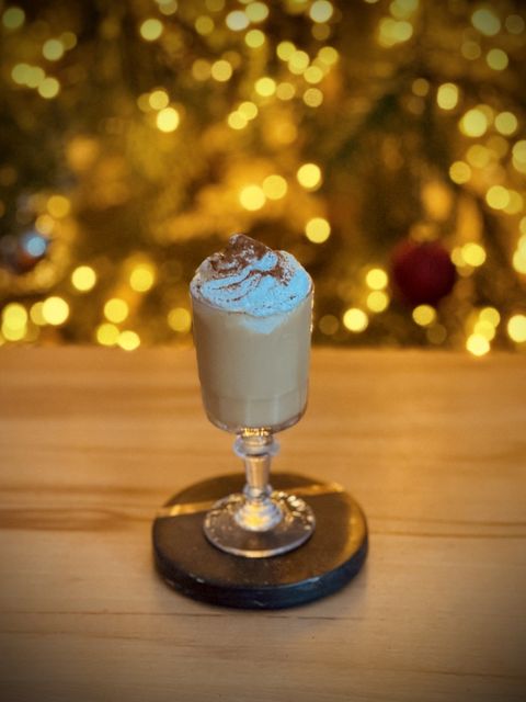 a creamy, light brown cocktail topped with whooped cream and garnished with grated cinnamon sitting in a crystal shot glass on a wooden table in front of a lit Christmas tree