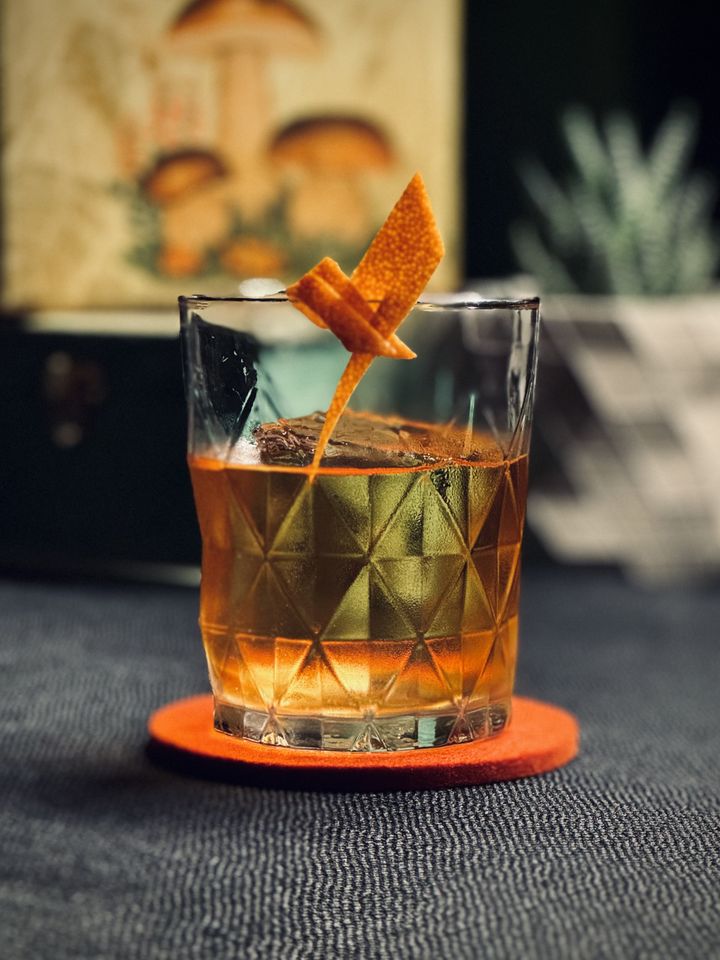 a light red cocktail with orange twist and big rock of clear ice in a geometric glass sitting on a red felt coaster in front of a plant and tea boxes