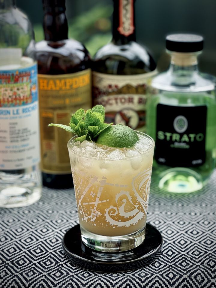 an off-white milky cocktail with mint bouquet and lime shell garnish, sitting on a black coaster and black and white patterned tablecloth with bottles of Clairin, Hampden, Doctor Bird, and Strato pistachio distillate in the background
