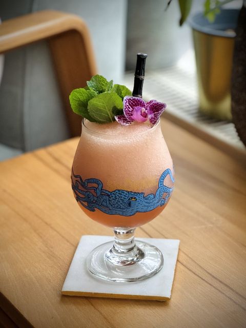 a salmon or light orange cocktail with mint bouquet and purple orchid garnish and a surfside sips realistic black bamboo glass straw in a Jester King Snörkel glass with a blue octopus on the front