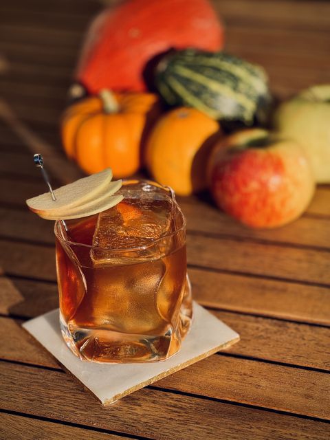 a sunlit, amber brown cocktail with apple slice fan garnish and big rock of clear ice in a warped hand-blown glass sitting on a white coaster on a wooden table with pumpkins and apples in the background