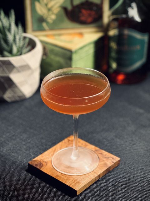 a brown cocktail in a chilled coupe glass sitting on an olive wood coaster in front of some bottles and a potted plant