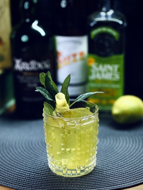 a bright yellow cocktail with large rock of clear ice, lemon peel bloom, and sage leaf sprig garnish sitting on a mat with bottles and a lemon in the background
