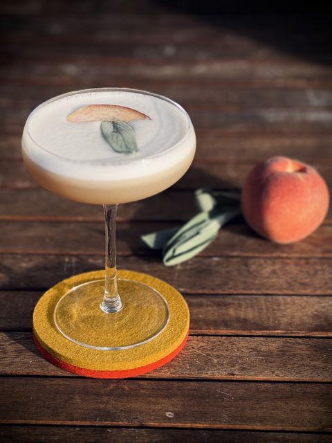 a light yellow cocktail with solid head of foam garnished with peach slice and sage leaf in a coupe glass sitting on a wooden table with a whole peach and group of sage leaves nearby
