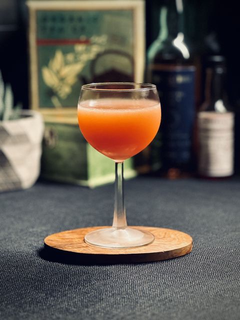 a bright red cloudy cocktail in a chilled coupe sitting on an olive wood coaster. In the background are bottles and a potted plant.