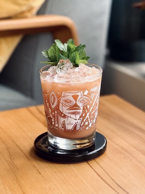 a creamy pink cocktail with mint bouquet garnish sitting on a black ceramic coaster on a wooden table with a chair and yellow pillow in the background