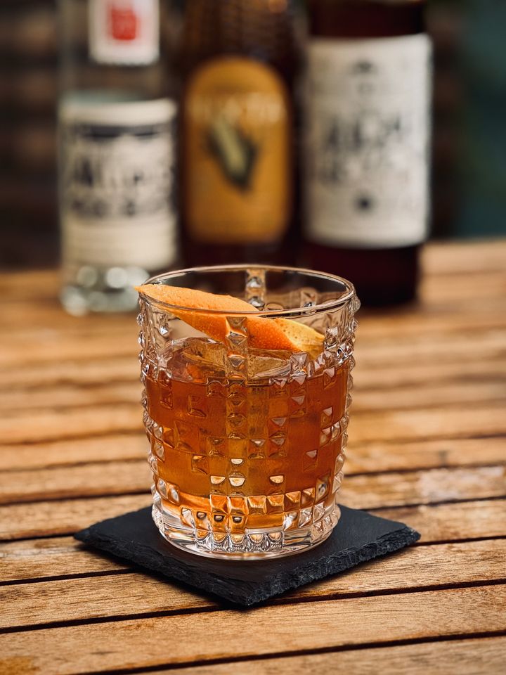 a dark amber brownish cocktail with orange peel garnish and big rock of clear ice sitting on a black coaster on a wooden table. In the background are bottles of Alipus mezcal, Nixta Elote liqueur, and Ancho Reyes chili liqueur