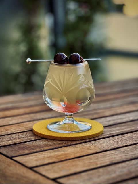 a light yellow cocktail with maraschino cherry garnish in an etched crystal brandy snifter sitting on a yellow felt coaster on a wooden table