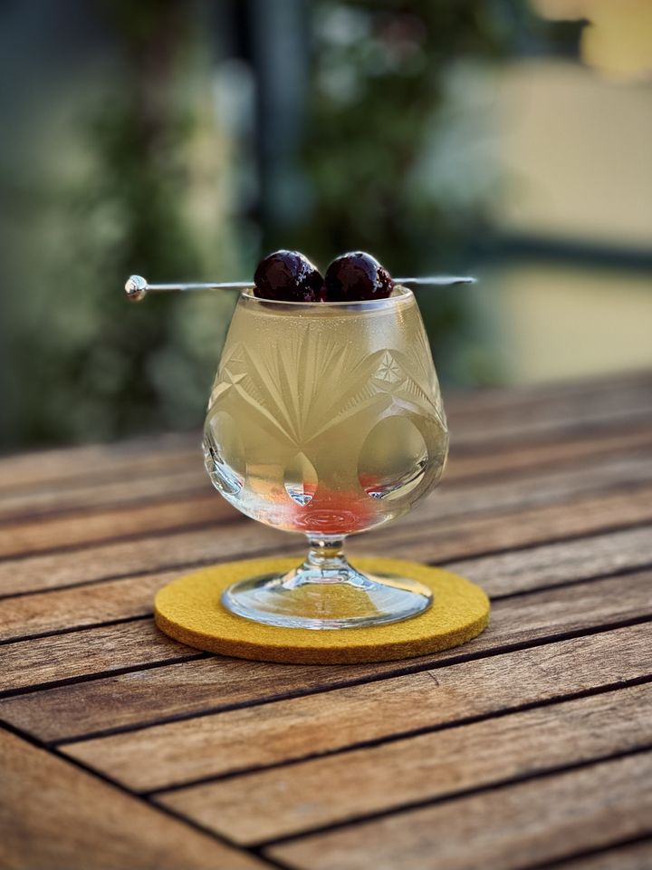 a light yellow cocktail with maraschino cherry garnish in an etched crystal brandy snifter sitting on a yellow felt coaster on a wooden table