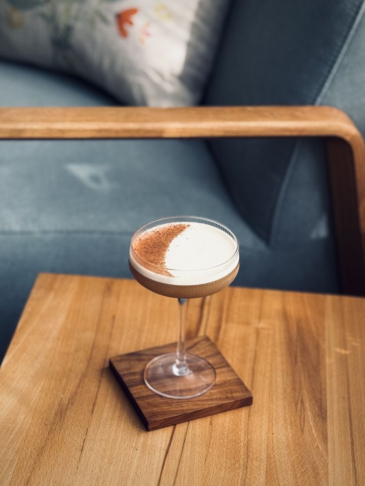 a creamy brown cocktail in a coupe glass with white foam top garnished with chocolate powder in a crescent moon shape sitting on a wooden table with a chair in the background