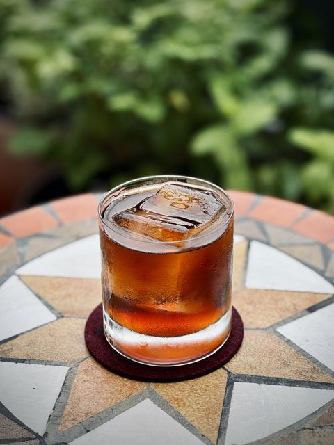a dark brown cocktail with big rock of clear ice sitting in a table with plants in the background
