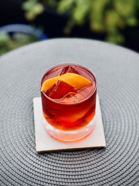 bright red cocktail with big rock of clear ice and orange twist garnish sitting on a white coaster with plants in the background