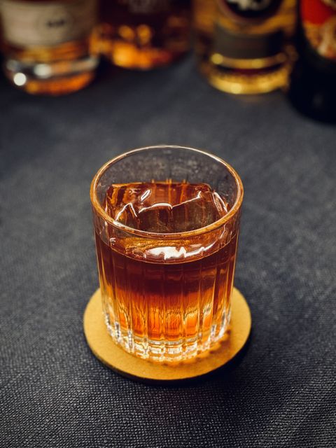 a clear brown cocktail with big rock of clear ice and no garnish sitting on a yellow coaster on a black surface with bottles in the background