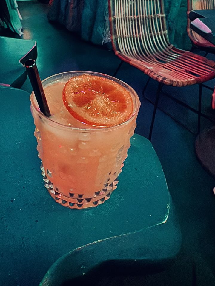 a light orange cocktail with black metal straw and candied orange garnish in a Nachtmann studded crystal glass sitting on a table at Mothership in San Diego with a wicker chair in the background