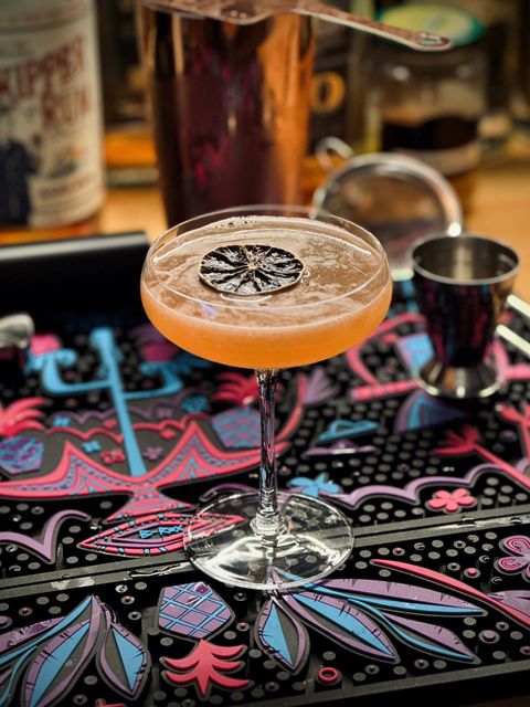 a light orange cocktail in a couple glass with dehydrated lime garnish floating on top. it's sitting on an Art of B-Rex bar mat with bartending equipment in the background