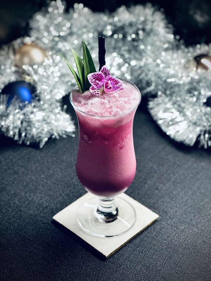 a bright pink cocktail with Surfside Sips black dragon glass straw, pineapple frondsm and purple orchid garnish in a hurricane glass sitting in front of silver garland and glass ball christmas ornaments