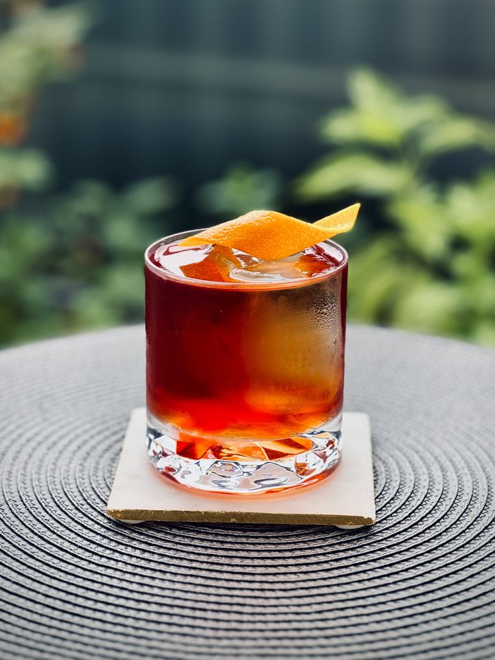 dark red cocktail with big rock of clear ice and orange twist garnish sitting on a circular mat with plants in the background