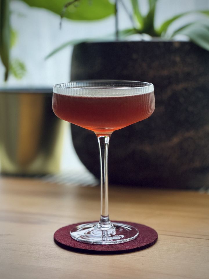a brown-red cocktail in a ribbed coupe class sitting on a purple felt coaster on a wooden table