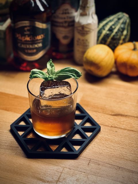 a dark brown cocktail with big rock of clear ice and basil sprig garnish sitting on a rubber lattice coaster with bottles and pumpkins in the background