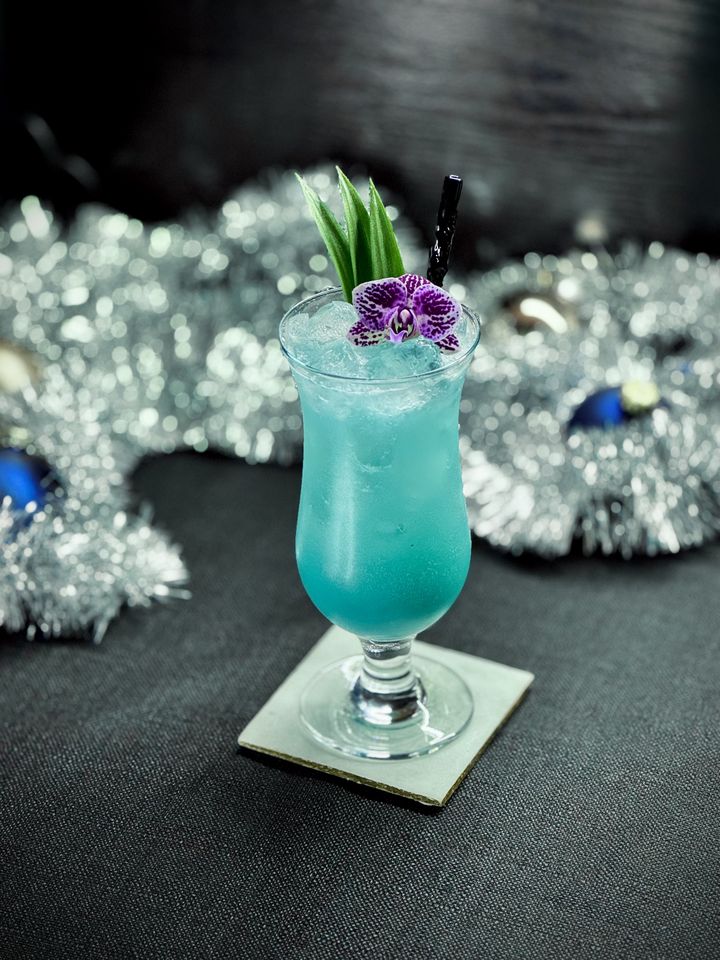 a bright blue cocktail with Surfside Sips black graf on glass straw, pineapple frond and purple orchid garnish in a hurricane glass with silver garland and glass ball christmas ornaments in the background