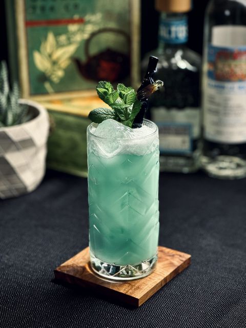 a creamy blue cocktail in a collins glass with a mint bouquet garnish and Surfside Sips realistic black bamboo straw with Ferdinand the Frog attached. In the background is a tea box and some bottles