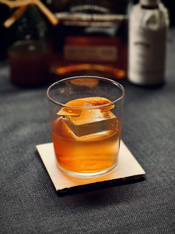 a golden brown cocktail with orange twist garnish and big rock of clear ice sitting on a white coaster in front of bottles
